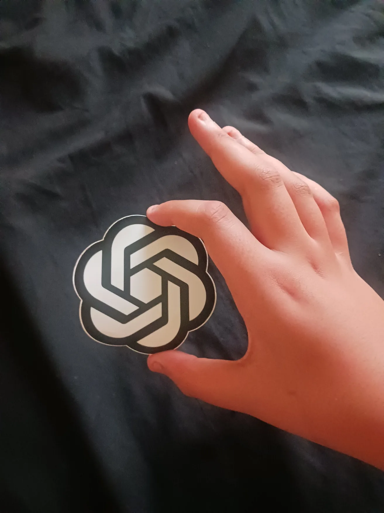 Mão segurando um adesivo com o símbolo da OpenAI sobre um tecido escuro